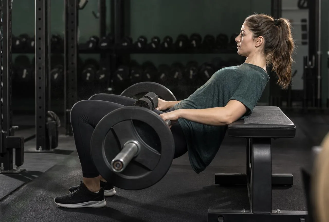 Mulher executando hip thrust com barra apoiada no quadril, costas apoiadas no banco e coluna neutra